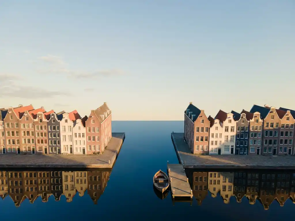 Aerial view of Amsterdam canal houses with terracotta facades reflected in still blue water, small wooden boat moored at a quiet dock in golden morning light.