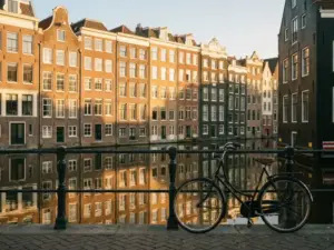 Amsterdamse grachtenpanden met terracotta gevels en grote ramen, fiets tegen ijzeren reling, weerspiegeling in kalm water, gouden middaglicht.
