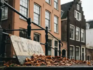 Verouderd Te Koop-bord voor een Amsterdams grachtenpand met warme bakstenen gevel en herfstbladeren aan de voet.