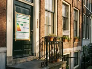 Energielabel certificaat op de voordeur van een Amsterdams grachtenpand met terracotta gevel en te koop bord.
