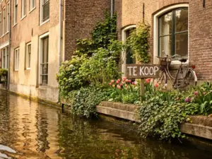 For Sale sign in front garden of Amsterdam canal house with bicycle against brick facade, golden spring light reflecting on water.