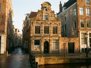 Historic Amsterdam canal house with ornate gabled facade, brick detailing, and a subtle for-sale sign beside the arched door at golden hour.