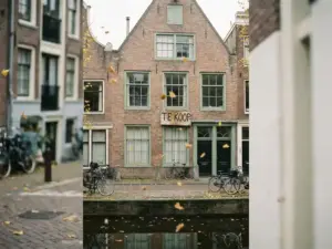 Amsterdam canal house with a weathered "Te Koop" for-sale sign, autumn leaves drifting past aged brick facade in soft overcast light.