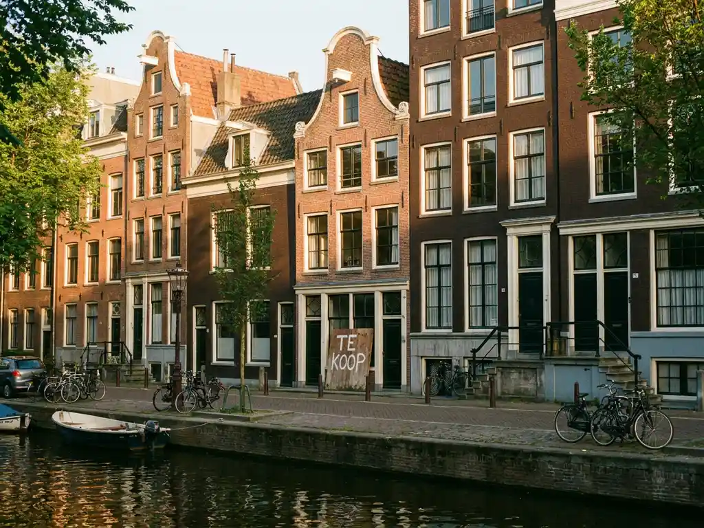 Dutch brick townhouse on tree-lined Amsterdam canal street with golden afternoon light and a "te koop" for-sale sign near the entrance.
