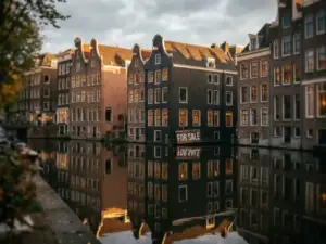 Amsterdam canal houses with a For Sale sign against brick facade, golden autumn light on gabled rooftops reflected in still water.