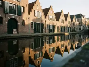Amsterdam canal houses with stepped gable rooftops reflected in still water, golden afternoon light on brick facades, for sale sign near arched doorway.