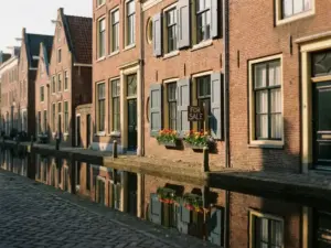 Amsterdam canal houses with tulip window box and "for sale" sign reflected in still water, bathed in warm afternoon light.